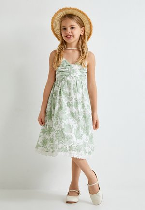 Robe fleurie verte avec des bordures en dentelle blanche, présentant un buste froncé et des bretelles spaghetti. Associée à un chapeau en paille et des chaussures blanches.
