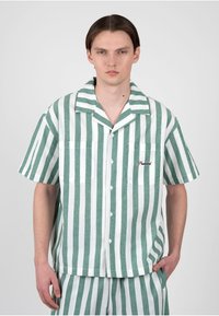 Jeune homme portant une chemise à boutons à rayures verticales vertes et blanches à manches courtes avec un short assorti, debout devant un fond uni.