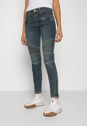 Person iført mørkeblå motorcykeljeans med ribbede knæpaneler, lynlåselementer, hvide sokker og hvide sneakers med brune såler.