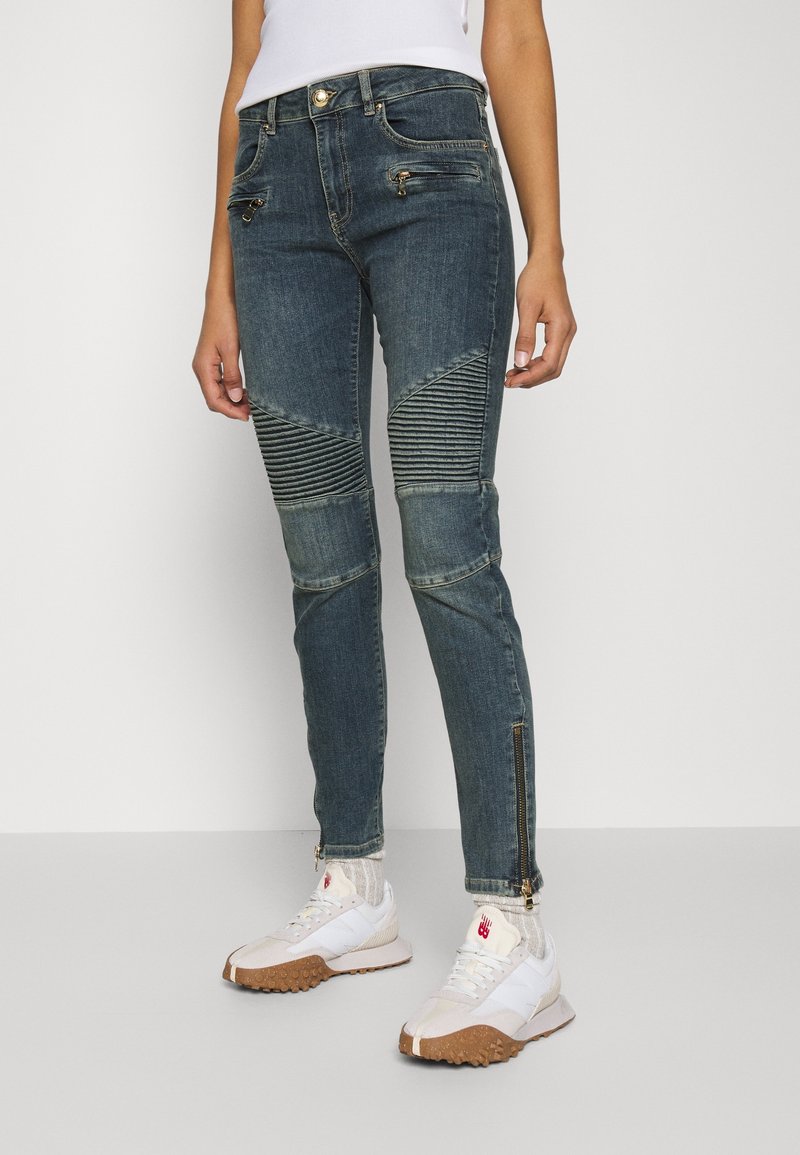 Personne portant un jean style motard bleu marine avec des panneaux côtelés aux genoux, des détails à fermeture éclair, des chaussettes blanches et des baskets blanches à semelles marron.