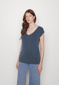 Camiseta azul escura com decote em V, mangas curtas dobradas, tecido leve, corte justo e textura suave. Usada com jeans de cintura alta.