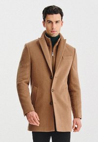 Brązowy wełniany płaszcz o wyciąganym kołnierzu, z dwoma guzikami i bocznymi kieszeniami. Nosi się go na swetrze z wysokim kołnierzem, co zapewnia dopasowany krój.