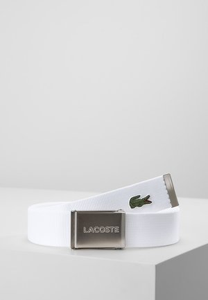 Ceinture en tissu blanc avec boucle en métal argenté gravée "LACOSTE" et logo crocodile vert brodé sur l'extrémité de la ceinture, présentée sur une surface blanche.