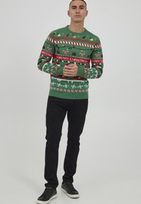 Grüner Pullover mit festlichen Mustern, dem Schriftzug "FROHE WEIHNACHTEN" und ikonischen Feiertagsmotiven. Kombiniert mit schwarzen Hosen und dunklen Sneakern.