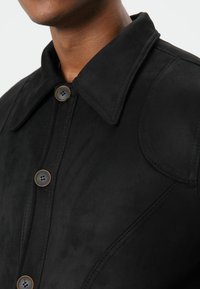 Zwarte suède jas met een puntige kraag, twee bruine knopen en gedetailleerde stiksels aan de voorkant en schouders.