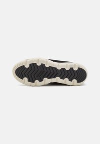 Suela de zapatilla deportiva negra con una entresuela blanca, que presenta un patrón de banda de rodadura en zigzag para mejorar la tracción. El nombre de la marca "SOREL" es visible en la suela.