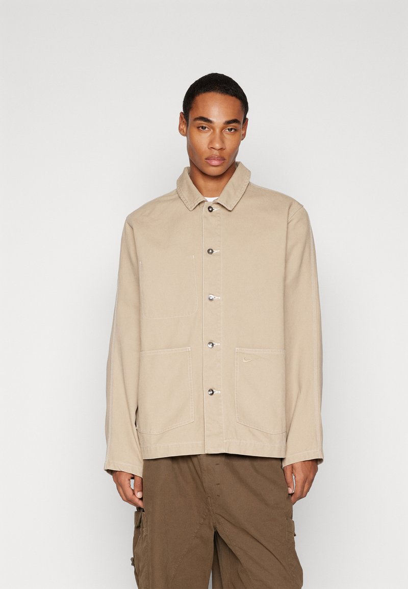Chaqueta de lienzo beige con cuello, cierre de botones, dos bolsillos frontales y detalle de logo sutil. Tejido texturizado con costuras visibles.
