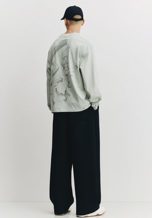 Camicia verde chiaro a maniche lunghe con stampa grafica sul retro, abbinata a pantaloni neri larghi e un berretto nero, creando un look minimalista.