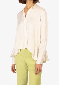 Camicia di seta crema con colletto, chiusura a bottoni e orlo svasato. Abbinata a pantaloni verde chiaro. Texture liscia.