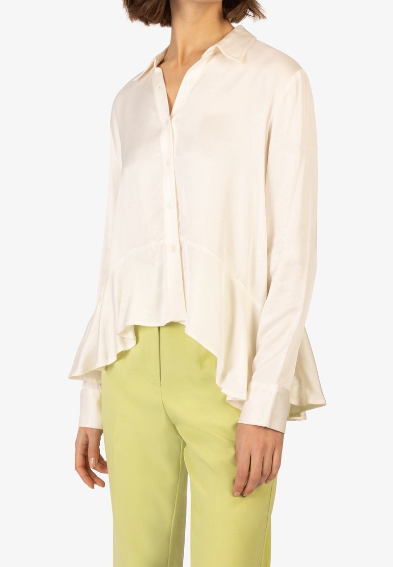 Camicia di seta crema con colletto, chiusura a bottoni e orlo svasato. Abbinata a pantaloni verde chiaro. Texture liscia.