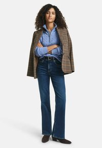 Brauner karierten Blazer über einem hellblauen Strickpullover, kombiniert mit dunkelblauen Schlagjeans und braunen Schuhen, die Arme verschränkt.