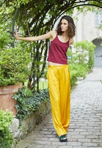 Débardeur bordeaux associé à un pantalon large plissé jaune vif. Le modèle porte des sandales noires avec des accents verts, devant un décor de jardin.
