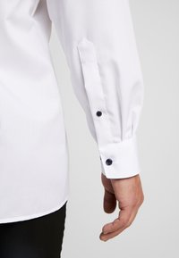 Weißes Langarmhemd mit geknöpften Manschetten. Verfügt über zwei dunkle Knöpfe, schmale Passform und glatte Stoffoberfläche. Die Manschetten haben einen spitzen Schlitz.