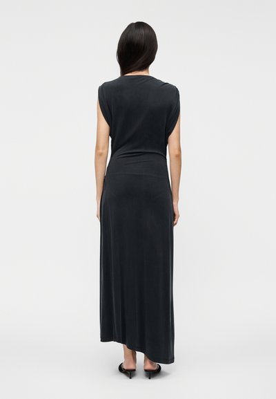 Woman with long dark hair wearing a sleeveless black maxi dress and black heeled sandals, standing against a plain white background.