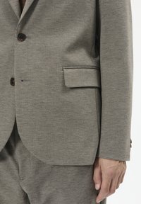 Blazer gri deschis în modele herringbone, cu un singur buzunar pe piept, două butoane și accente de butoane maro. Materialul are un finisaj neted și texturat.