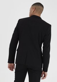 Hombre con cabello corto que lleva aretes de aro dorados y un blazer negro ajustado con pantalones, visto desde atrás contra un fondo liso.