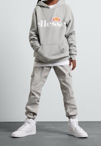 Grauer Kapuzenpullover mit weißem "ellesse"-Logo und orangefarbenem Akzent. Helle graue Cargo-Hosen und weiße High-Top-Sneaker runden das Outfit ab.