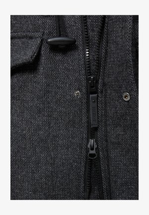 Grijs gestructureerde jasstof met een zwarte rits en twee drukknoopjes. Elastisch koord met plastic trekker bij de kraag.