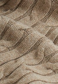 Tissu en laine beige tricoté présentant une texture ondulée avec des motifs câblés en relief sur toute la surface, offrant une apparence chaleureuse et tactile.