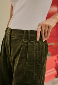 Pantaloni din corduroy de culoare verde măslinie, cu pliseuri și buzunare laterale vizibile. Materialul are un model texturat de grilă. Se poartă un top alb, ajustat.