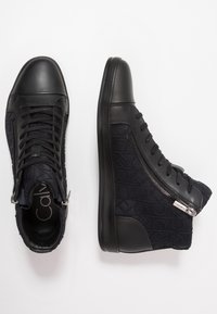 Černé vysoké tenisky s hladkou koženou špičkou, texturovanou tmavou látkou s logem a bočním zipem. Černá gumová podešev.