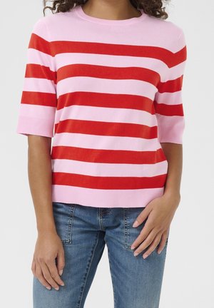 Persoon draagt een roze en rood horizontaal gestreepte korte mouwen trui, gecombineerd met blauwe spijkerbroek, handen ontspannen langs het lichaam.