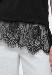 Bordure en dentelle noire avec des motifs floraux sur l'ourlet d'un haut noir porté sur un pantalon blanc, avec une main reposant sur le côté.