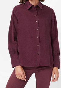 Chemise en velours côtelé bordeaux avec un col, des manches longues et des boutons en métal argenté sur le devant. Elle présente un tissu texturé et une coupe décontractée.