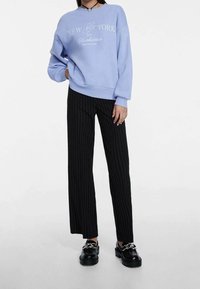 Lichtblauwe sweatshirt met witte tekst, relaxte pasvorm, geribbelde manchetten, gecombineerd met zwarte broek met fijne streep en zwarte schoenen met kettingdetail.