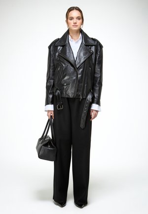 Donna che indossa una giacca da motociclista in pelle nera, camicia bianca, pantaloni larghi neri, tiene una borsa nera in mano, in piedi davanti a uno sfondo bianco.