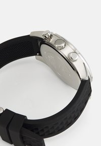 Reloj cronógrafo de acero inoxidable plateado con una correa de goma negra que presenta un patrón de diamante texturizado y dos pulsadores en el lado.