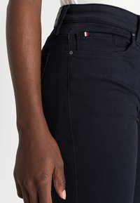 Jeans de mezclilla azul oscuro de cintura alta, con un pequeño detalle de bandera roja, blanca y azul en el bolsillo y sutilezas en la costura.