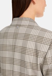 Kariertes Hemd mit grauem und beigem Muster und markanten Linien. Das Rückendetail umfasst einen Knopf und eine glatte Stofftextur.