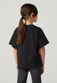 Laps, kellel on pikad pruunid juuksed, seotud hobusesabas, kannab lahtist musta T-särki ja tumedaid teksaseid, selg pöördunud ühtlase tausta poole.