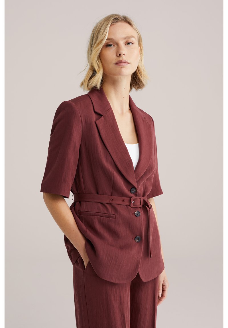 Mujer con cabello corto y rubio, vistiendo un blazer color burdeos con cinturón y botones, y pantalones a juego, sobre un fondo liso.
