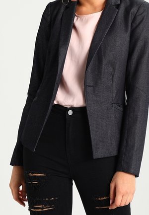 Veste cintrée gris foncé avec revers crantés, tissu texturé et deux poches latérales, associée à un blouse rose clair et un pantalon en jean noir déchiré.