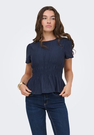 Mujer con cabello largo y ondulado, vestida con una blusa fruncida de manga corta azul marino y vaqueros azul oscuro sobre un fondo liso y claro.
