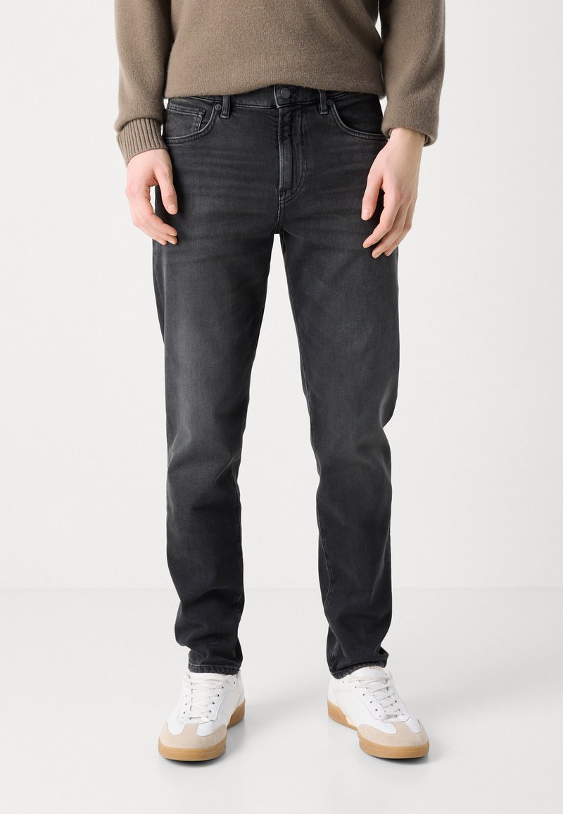 Black denim jeans with a slim fit, featuring five pockets, subtle whiskering on the thighs, paired with white sneakers.