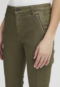 Pantalons cargo vert olive en tissu texturé, avec des poches zippées à l'avant et une coupe ajustée. Gros plan sur la taille et le placement des mains.