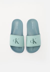 Helle blaue Slide-Sandalen mit einem strukturierten Riemen, auf dem das erhabene "cK"-Logo prangt. Die Sohlen sind in einem dunkleren Blau gehalten und haben eine glatte Oberfläche.