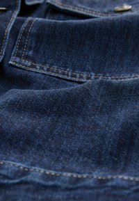 Mørkeblå denimstof med synlig tekstur og syninger. Har en brystlomme med en metalknap som accent.