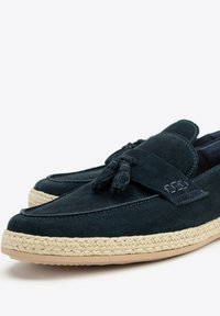 Dunkelblaue Wildlederslipper mit Quastenakzenten, ausgestattet mit einer geflochtenen Jutesohle und glatter Textur. Runde Zehenpartie und flache Absatzgestaltung.