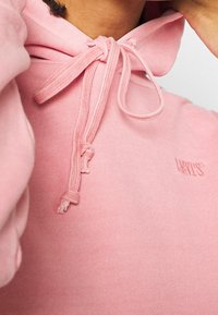 Vaaleanpunainen huppari pehmeästä puuvillasta. Varustettu säädettävällä kiristysnyörillä ja hupulla, resoripäillä sekä pienellä kirjotulla logolla rinnassa.