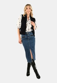 Gonna a tubino in denim con spacco frontale, abbinata a una camicetta bianca con dettagli blu navy e un gilet nero in maglia, completata da stivali neri al ginocchio.
