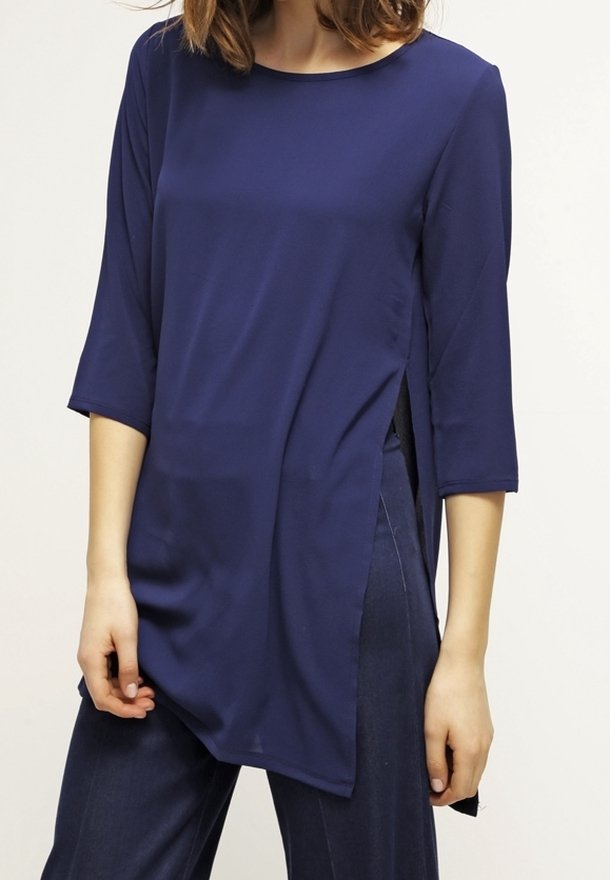 Femme portant une tunique asymétrique bleu marine avec des manches trois-quarts et des fentes latérales, associée à un pantalon bleu foncé.