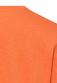 Zbliżenie na jednolitą pomarańczową tkaninę z widocznym przeszyciem wzdłuż górnej krawędzi, prawdopodobnie element ubrania takiego jak koszulka lub bluza.