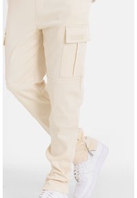 Beige cargo broeken met patch pockets, een aansluitende pasvorm en een zachte textuur, gecombineerd met witte sneakers en een ritssluiting op de broeken.