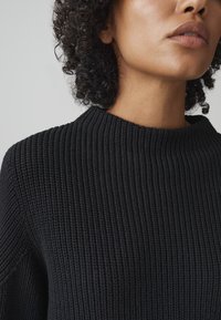 Maglione nero lavorato a maglia con texture a coste, scollatura rotonda e maniche aderenti. Vista ravvicinata che mette in risalto i dettagli e la struttura del tessuto.