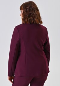Frau mit schulterlangem, welligem braunem Haar, die ein enges, dunkelviolettes Blazer und passende Hose trägt, mit dem Rücken zu einem schlichten Hintergrund gewandt.