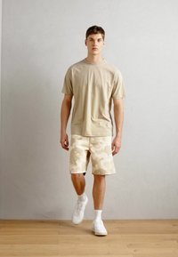 Beige t-shirt and cream camo shorts, with white sneakers. The fabric appears soft; the shorts feature a subtle, tonal pattern.